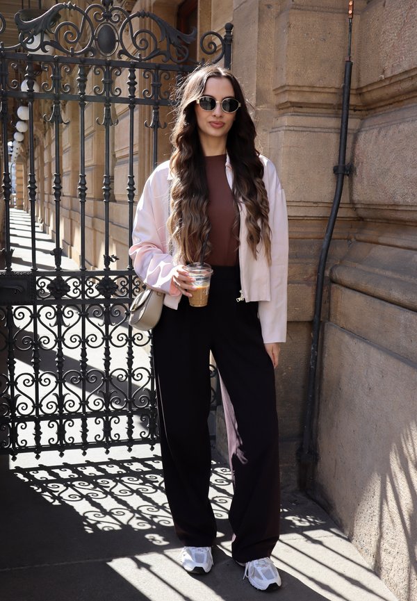 Femme aux longs cheveux ondulés portant des lunettes de soleil, une veste légère, un haut marron, un pantalon noir et des baskets blanches, tenant un café glacé près d'une grille en fer décorative.