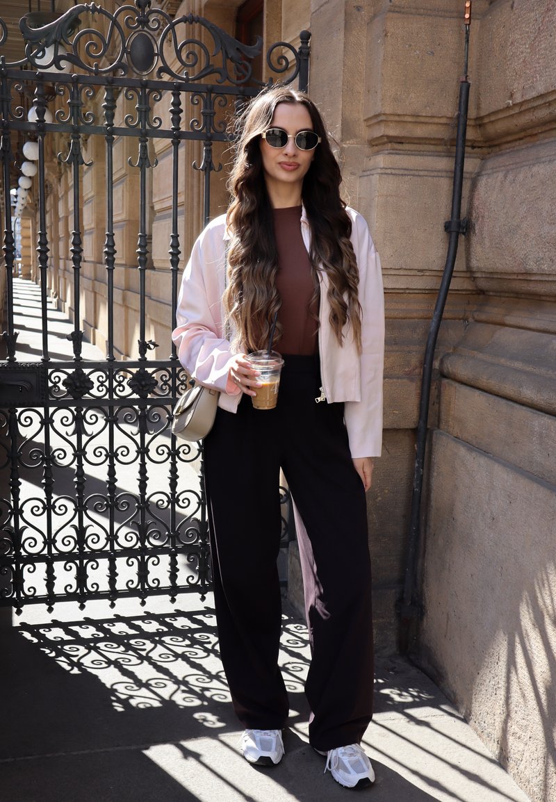 Femme aux longs cheveux ondulés portant des lunettes de soleil, une veste légère, un haut marron, un pantalon noir et des baskets blanches, tenant un café glacé près d'une grille en fer ornée.