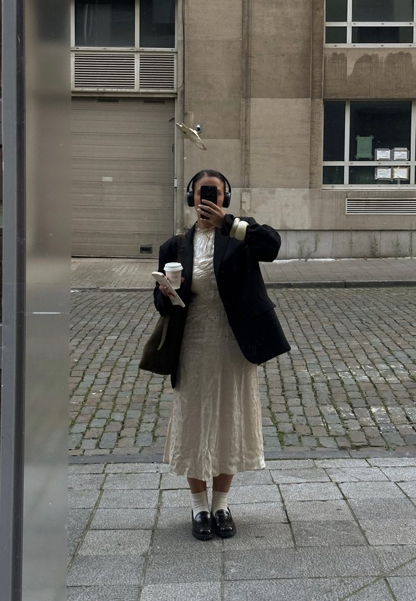 Blazer noir sur une robe midi légère et texturée ; tenant une tasse de café et un téléphone ; portant des écouteurs ; chaussettes montantes et mocassins.
