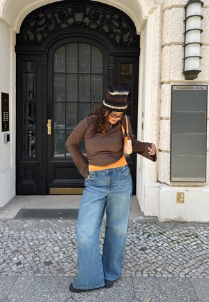 Chemise marron à manches longues par-dessus un haut orange, jean large bleu, et un bonnet en tricot noir et marron à rayures. Tenue décontractée avec des éléments texturés.
