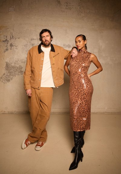 Hombre con chaqueta y pantalones color topo y zapatillas blancas se apoya en una mujer que lleva un vestido brillante con estampado de leopardo y botas negras hasta la rodilla, apoyados contra una pared texturizada.