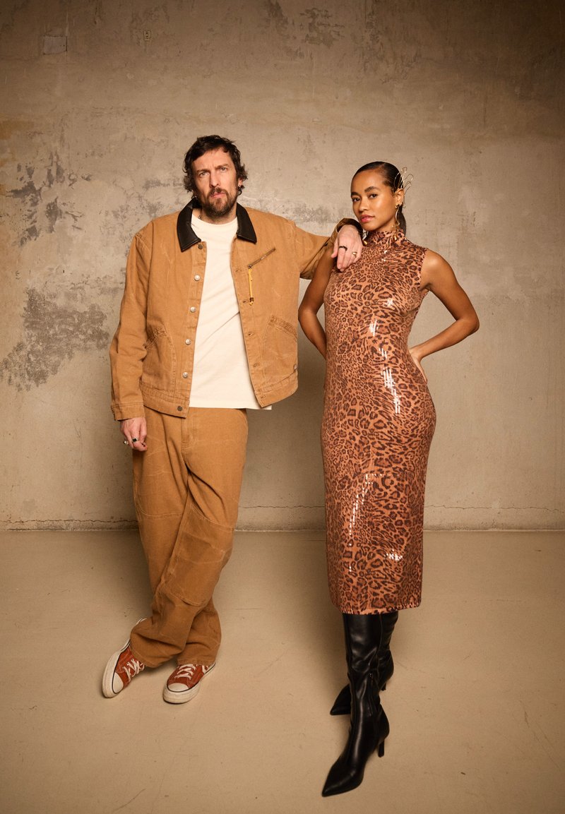 Bărbat îmbrăcat într-o jachetă și pantaloni culoarea nisipului, cu adidași albi, se sprijină de o femeie care poartă o rochie sclipitoare cu imprimeu leopard și cizme negre până la genunchi, în fața unui perete texturat.