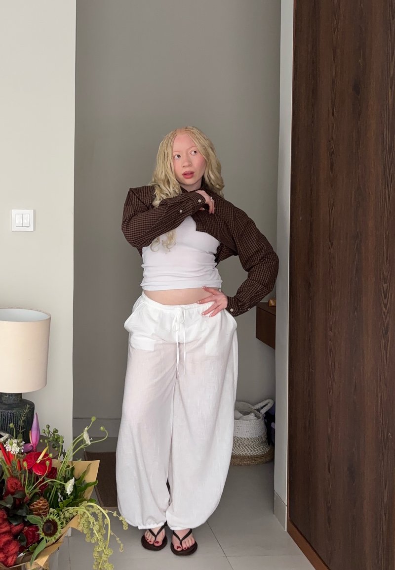 Person with long blonde hair wearing a cropped brown jacket, white tank top, loose white pants, and black flip-flops standing indoors near flowers.