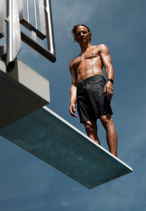 Trampolín con una superficie suave de color azul claro; una persona sin camiseta en bañador negro está en posición sobre él contra un cielo azul.