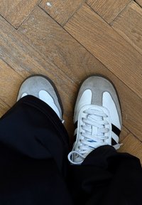 Baskets blanches et grises avec des bandes noires, dotées d'une tige en cuir et en daim, d'un bout rond et d'une semelle en caoutchouc texturée sur un sol en bois.