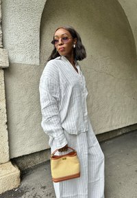 Camicia di lino grigio chiaro con righe verticali, dettaglio dei bottoni, abbinata a pantaloni a gamba larga a righe. Tiene una borsa intrecciata gialla con accenti marroni.