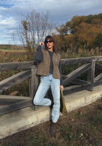 Colete de lã castanho sobre um suéter cinza, jeans azul claro e botas de tornozelo castanhas. A segurar uma bolsa verde-azeitona. Cerca de madeira e plantas ao fundo.