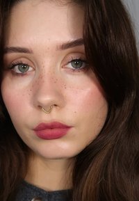 Närbild av ett ansikte med ljusbrunt, vågigt hår, gröna ögon, en guldfärgad septumring och matt rosa läppstift med synliga fregner på kinderna.