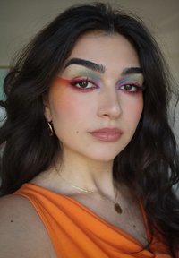 Porträtt av en person med långt, mörkt, vågigt hår. De har på sig färgglad ögonskugga i flera färger, guldörhängen med ringar och en orange topp.