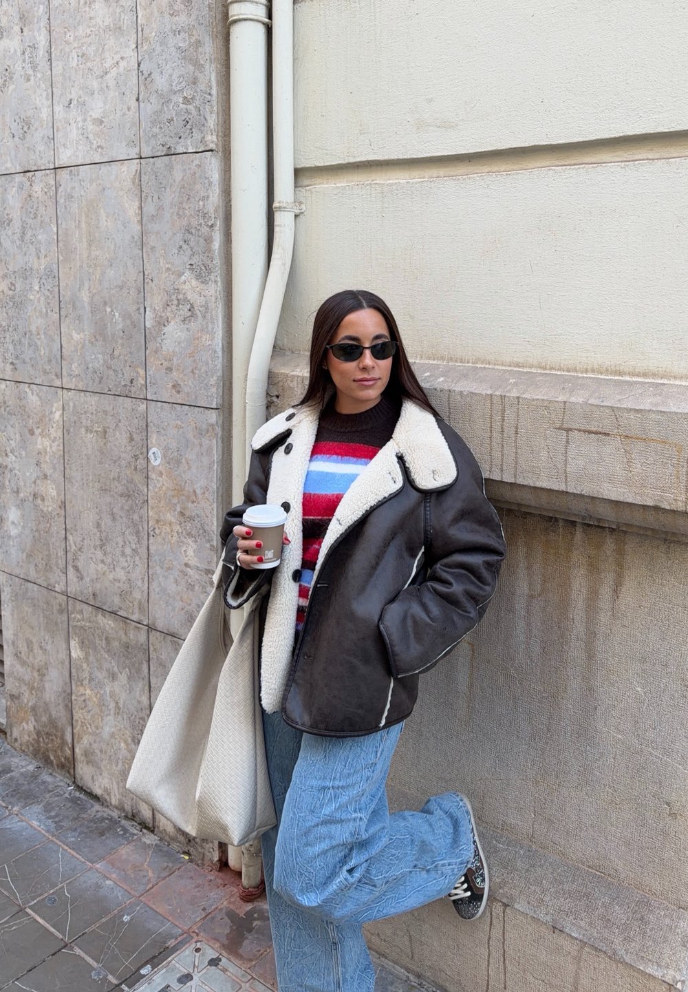 Femme portant une veste en shearling noire, un pull rayé, un jean bleu, des lunettes de soleil, tenant une tasse de café, appuyée contre un mur à un coin de rue urbain.
