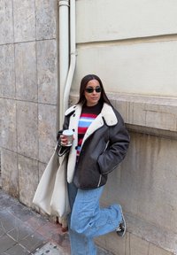 Femme portant une veste en shearling noire, un pull rayé, un jean bleu, des lunettes de soleil, tenant une tasse de café, appuyée contre un mur à un coin de rue urbain.