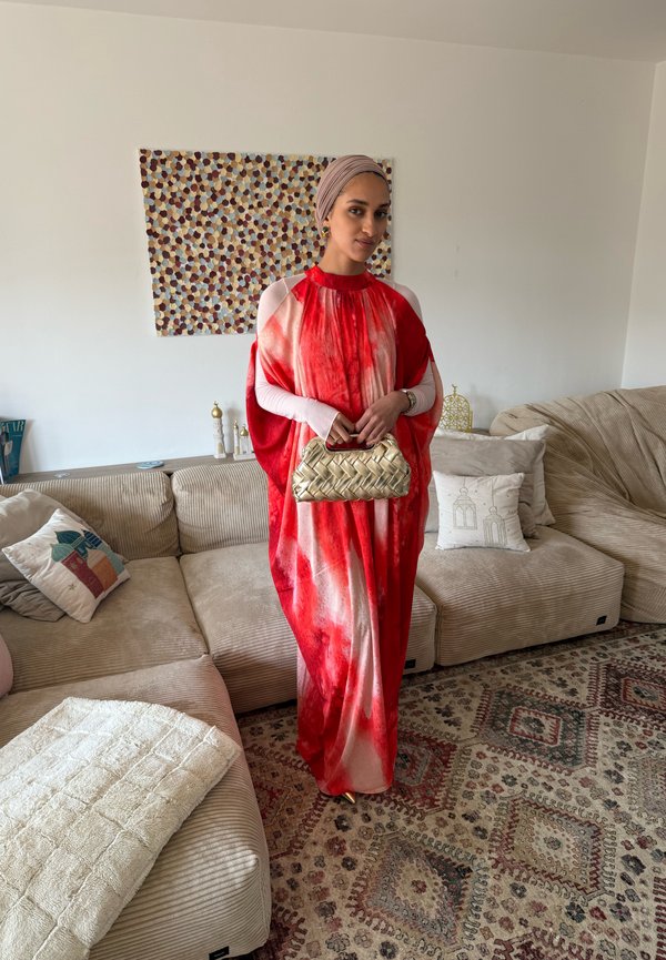 Ženska, oblečena v rdečo in belo obleko ter z glavo pokrito z ruto, drži zlato pleteno torbico in stoji v dnevni sobi z bež kavči in vzorčasto preprogo.