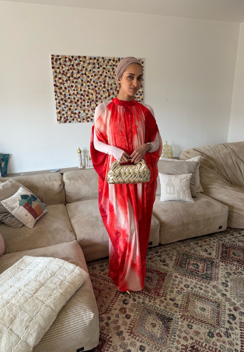 Frau trägt ein rotes und weißes Kleid und ein Kopftuch, hält eine gewebte goldene Handtasche und steht in einem Wohnzimmer mit beigen Sofas und gemustertem Teppich.