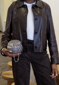Braune Lederjacke mit Kragendesign, Frontknöpfen und einer Tasche. Hält eine runde, metallische, silberne Perlenhandtasche mit einer Kettenriemen.