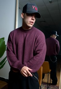 Burgunderfarbiger gerippter Pullover mit Rundhalsausschnitt, kombiniert mit schwarzen Hosen mit Kettendetails. Schwarze Mütze mit roter Aufschrift, innere Umgebung.