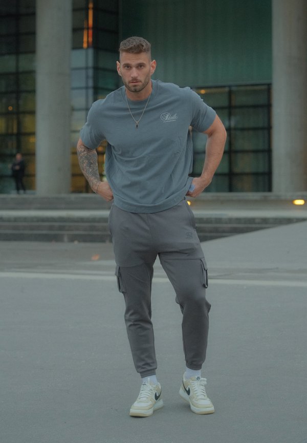Szürke póló logóval, szürke cargo nadrág és fehér sportcipő fekete részletekkel. Lazább szabású és diszkrét részletezéssel rendelkezik.