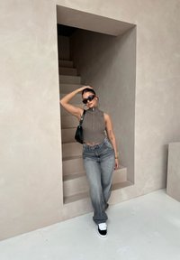 Kvinde i en tætsiddende brun zip-up trøje og højtaljede grå jeans, stående nær lyse trapper, iført sorte solbriller og sneakers.