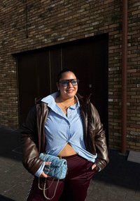 Brun läderjacka, ljusblå randig skjorta med öppen krage, vinröda byxor och en liten teal-färgad vadderad handväska med kedjeriem.