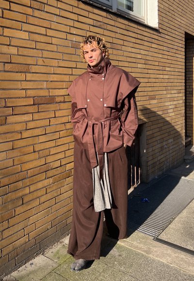 Persona con cabello rizado y rubio, vestida con una chaqueta marrón con cinturón y detalle de capa, y pantalones marrones anchos, de pie contra una pared de ladrillos bajo la luz del sol.