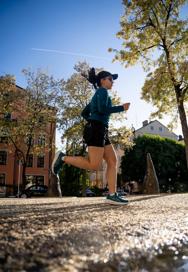 Biegowe buty w kolorze turkusowym, krótkie czarne spodenki sportowe oraz długi rękaw w tym samym kolorze. Okulary przeciwsłoneczne na nosie, jogging na fakturowanej nawierzchni z drzewami w pobliżu.
