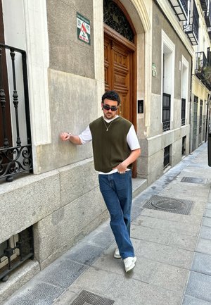 Suéter sin mangas color verde oliva sobre una camiseta blanca, combinado con jeans azules y zapatillas blancas, de pie contra una pared de piedra.