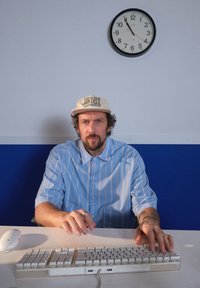 Valge arvutiklaviatuur ja hiir laual. Lokkis juustega inimene sinistes triibulistes särkides ja beeži mütsiga, käed klaviatuuril. Kell seinal.