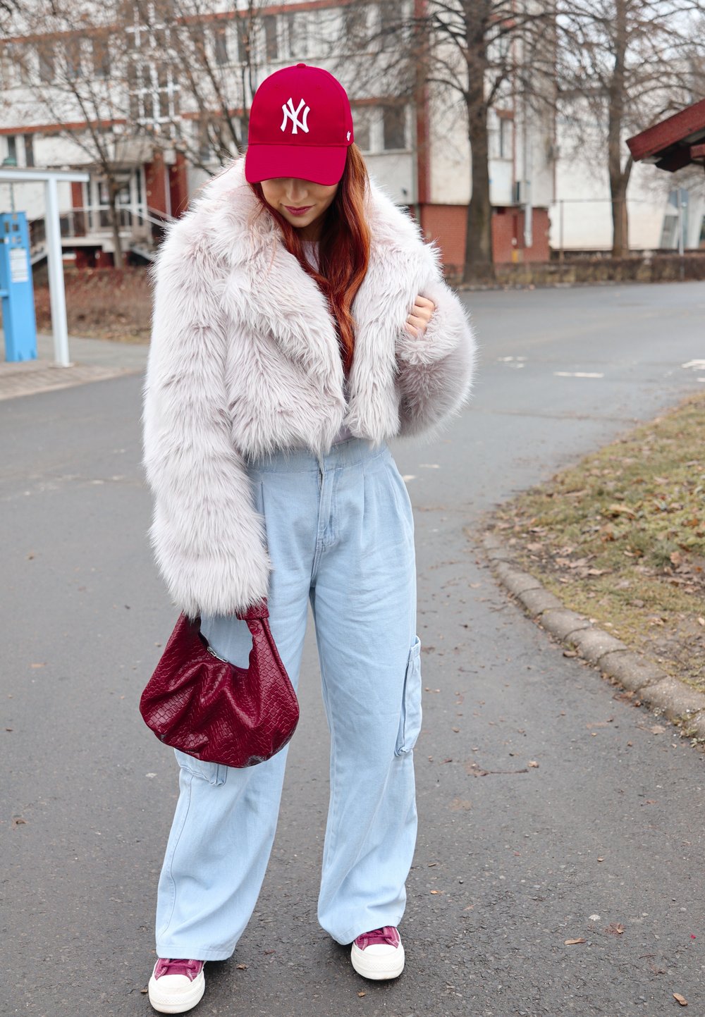 Ροζ ψεύτικο γουνάκι με κοντό κόψιμο, μπλε φαρδιές cargo παντελόνες, μπορντώ τσάντα χειρός, κόκκινο καπέλο baseball και λευκά αθλητικά παπούτσια με μπορντώ λεπτομέρειες.