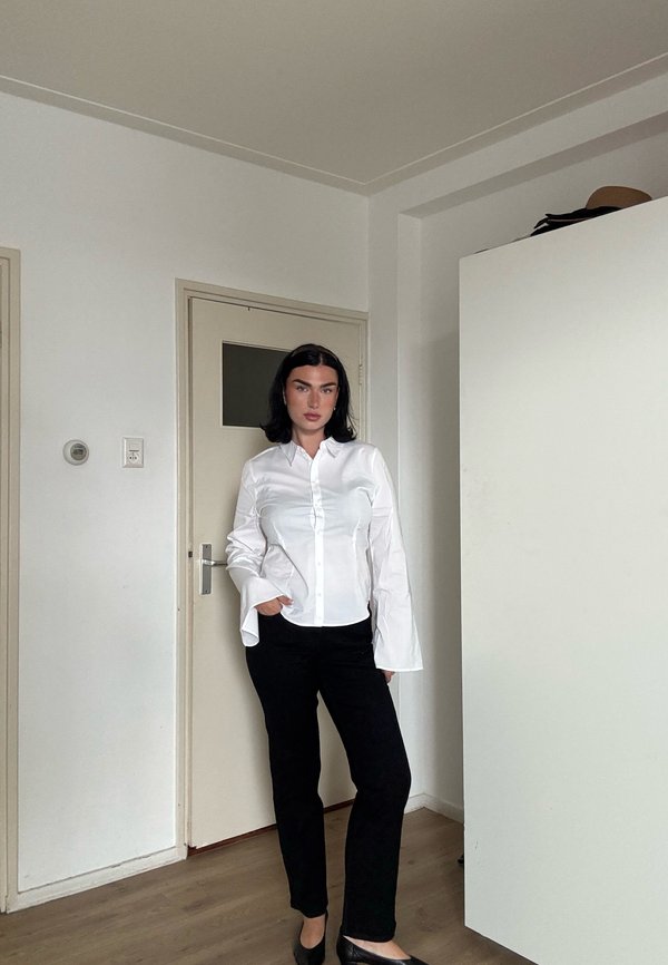Femme aux cheveux noirs portant une chemise blanche boutonnée et un pantalon noir, debout à l'intérieur près d'une porte beige et d'une armoire blanche.
