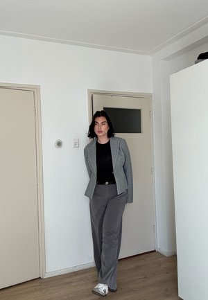Mujer de pie en el interior con blazer gris, camiseta negra, pantalones grises y zapatillas blancas frente a una pared blanca con puertas cerradas.