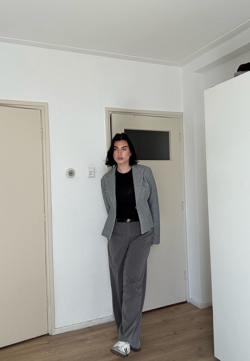 Femme debout à l'intérieur portant un blazer gris, un haut noir, un pantalon gris et des baskets blanches devant un mur blanc avec des portes fermées.