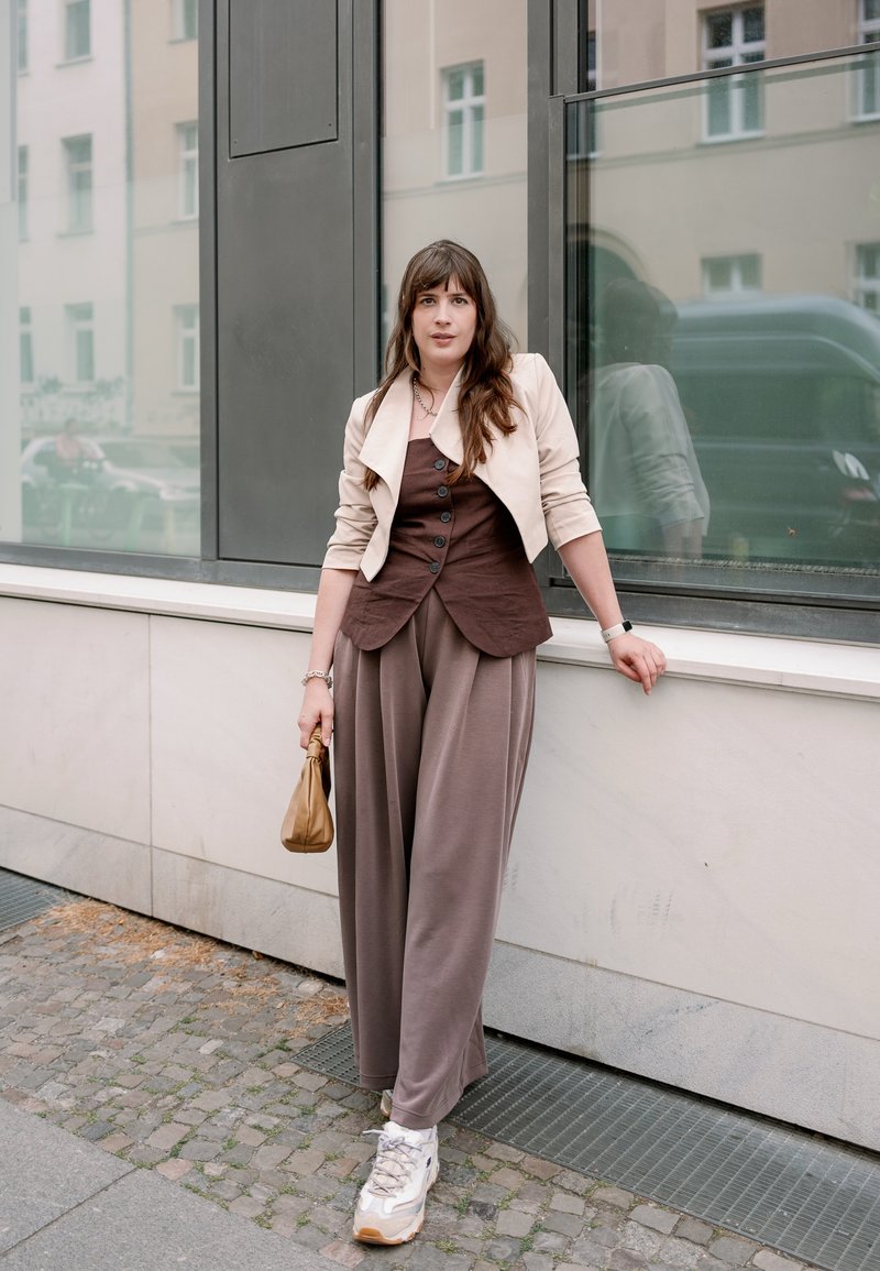 Jeune femme aux longs cheveux bruns appuyée contre une fenêtre d'un bâtiment, portant une veste beige, un top marron, un pantalon gris ample, des baskets blanches, et tenant un petit sac.