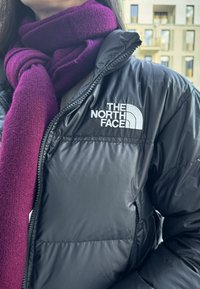 Schwarze Daunenjacke mit glänzender Oberfläche, versehen mit einem weißen Logo auf der linken Brust und einem dunkel-lila Schal, der um den Hals gewickelt ist.