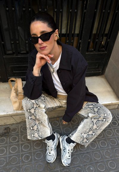 Mujer con gafas de sol negras, chaqueta negra, camisa blanca, pantalones con estampado de serpiente y zapatillas, sentada en un escalón con un bolso beige cerca.
