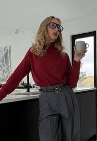 Roter Strickpullover mit hohem Kragen, kombiniert mit hoch taillierter grauer Plisseehose und braunem Gürtel. Hält eine hellgraue Tasse in der Hand.