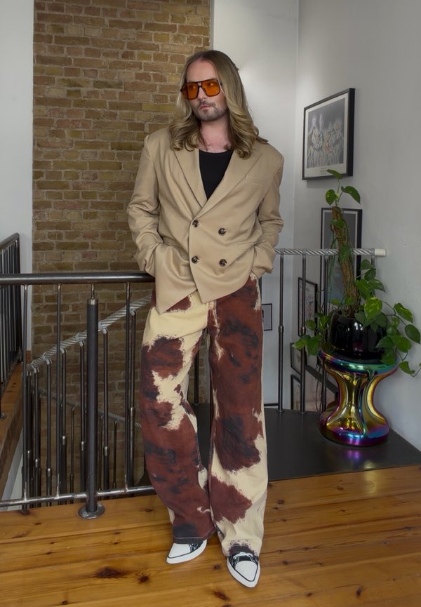 Personne aux longs cheveux portant des lunettes de soleil orange, un blazer beige, un pantalon large tie-dye et des chaussures blanches, se tient à l'intérieur près d'une rambarde et d'une plante.
