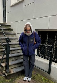 Veste polaire bleu marine avec une fermeture éclair et une poche poitrine, assortie à un pantalon gris et des chaussures de sport de couleur claire, debout sur des marches en pierre.