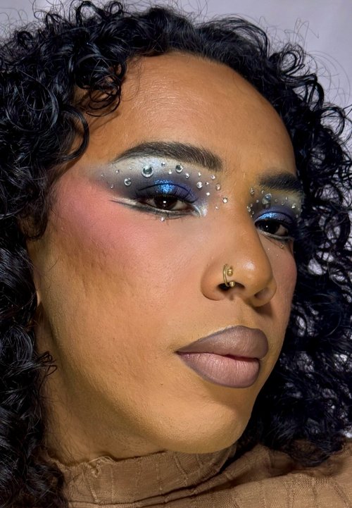 Person mit lockigem Haar, die blaue und silberne Lidschatten mit kleinen Strasssteinen trägt, neutralen Lippenstift und einen goldenen Nasenring.