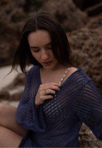 Pardesiu din croșetă albastru, cu țesătură liberă, mâneci lungi și decolteu adânc în formă de V. Elemente notabile includ o curea multicoloră și un inel pe mână.