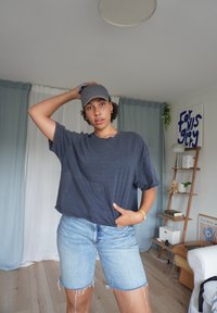 Tricou gri închis cu o croială lejeră, asortat cu pantaloni scurți din denim albastru deschis. Se poartă o șapcă gri, cu un logo brodat.