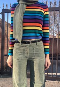 Άτομο που φοράει πολύχρωμη, μακρυμάνικη μπλούζα με ρίγες, παντελόνι κοτλέ χακί, μαύρη ζώνη και αποχρώσεις χακί κασκόλ, στέκεται έξω κοντά σε μια πύλη.