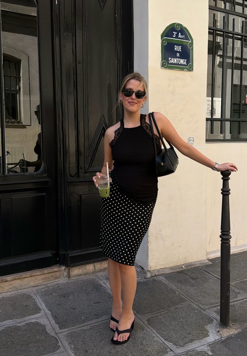 Femme en jupe noire à pois et haut, debout dans la rue Rue de Saintonge, tenant une boisson verte et s’appuyant sur un poteau noir.