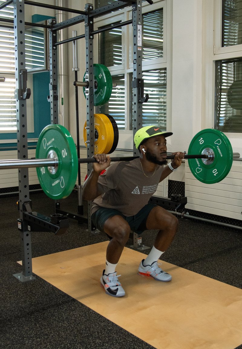 Homme portant une casquette et des écouteurs effectue un squat avec haltère dans une salle de sport, tenant une barre avec des disques de poids verts sur ses épaules.