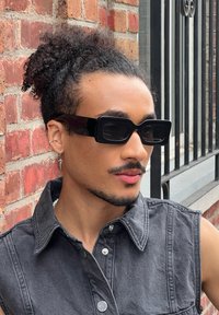 Czarne prostokątne okulary przeciwsłoneczne, teksturowana dżinsowa kamizelka, tło z ceglanej ściany, krótkie, kręcone włosy i srebrna kolczyk w kształcie koła.