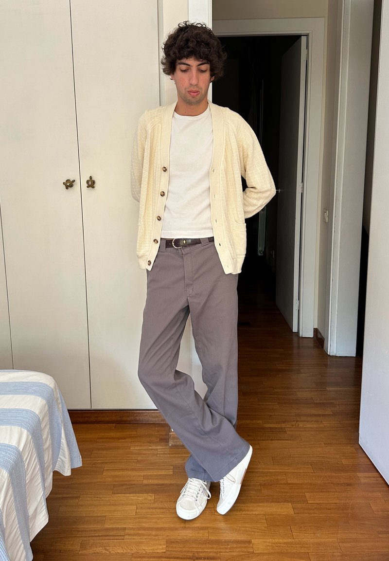 Junger Mann steht drinnen mit den Händen hinter dem Rücken, trägt eine beige Strickjacke, ein weißes T-Shirt, graue Hose und weiße Turnschuhe auf Holzboden.