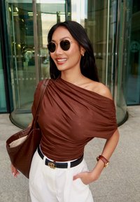 Rudos spalvos, nuo pečių nusmukusi palaidinė; baltos, aukštu liemeniu kelnės; juodas diržas su logotipu; ruda pintinė rankinė; apvalūs akiniai nuo saulės; glotnūs plaukai.