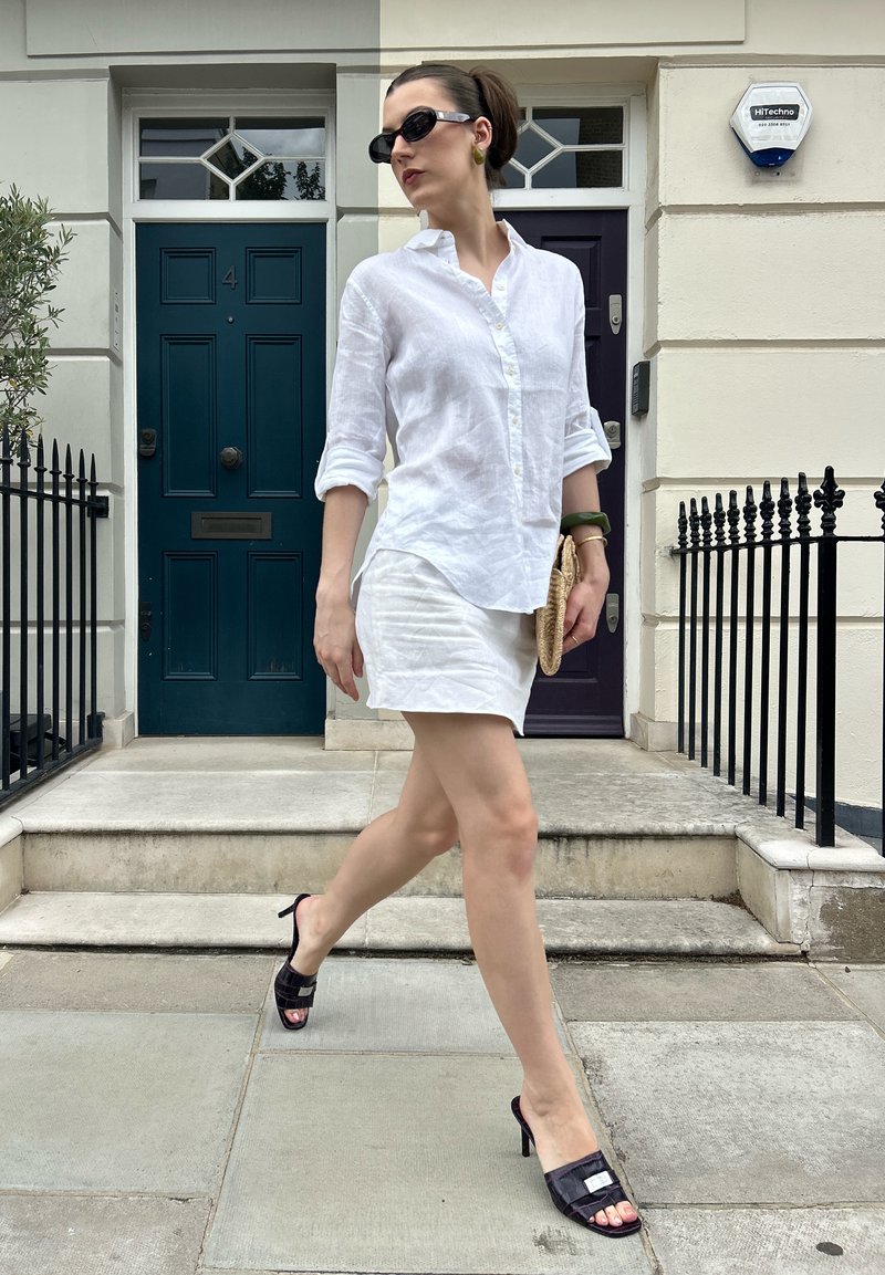 Femme en chemise blanche et jupe marchant devant des portes, portant des talons noirs, des lunettes de soleil, un bracelet vert et tenant une petite pochette tissée.