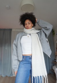 Grå kort zip-upp sweatshirt, vit lång halsduk, vit linne och blå jeans. Halsduken har fransar; slät textur överallt.