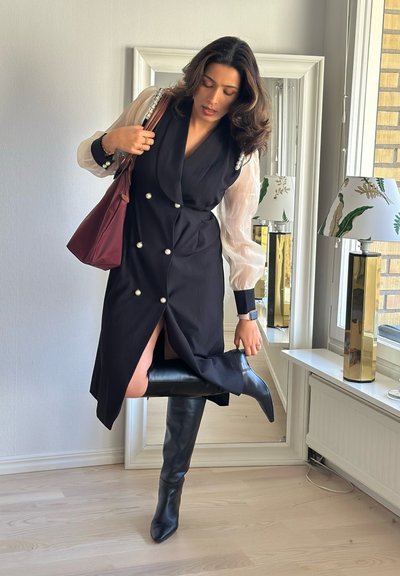 Vestido negro con escote en V profundo, diseño de doble botonadura y acentos de perlas. Las mangas blancas transparentes y las botas negras hasta el muslo completan el conjunto.