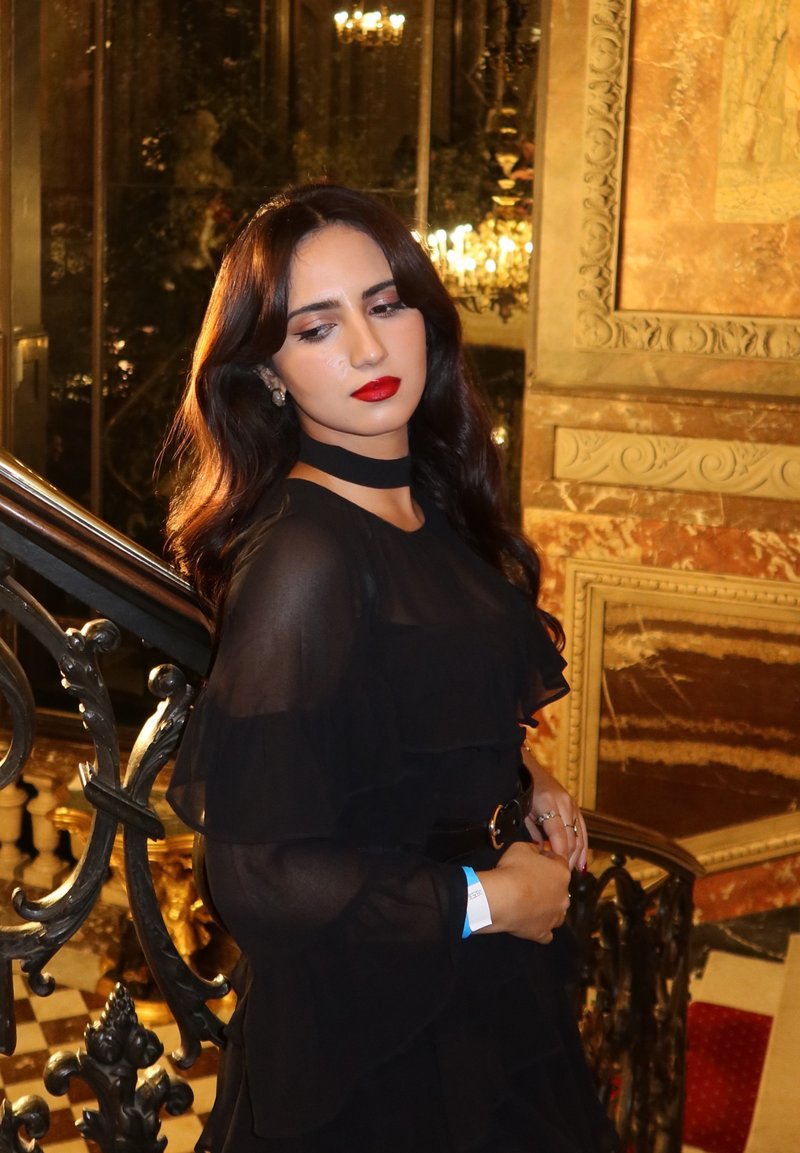 Robe en chiffon noire à plusieurs couches avec des volants ; cheveux longs et ondulés ; lèvres rouges ; debout près d'un escalier orné avec une balustrade en métal noir intricate.