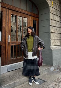 Brun skinnjakke i store størrelser, grønn strikkegenser, hvit bluse med rysjer, svart skjørt med prikker, grå sokker, hvite høye hæler, mørk burgunder håndveske.